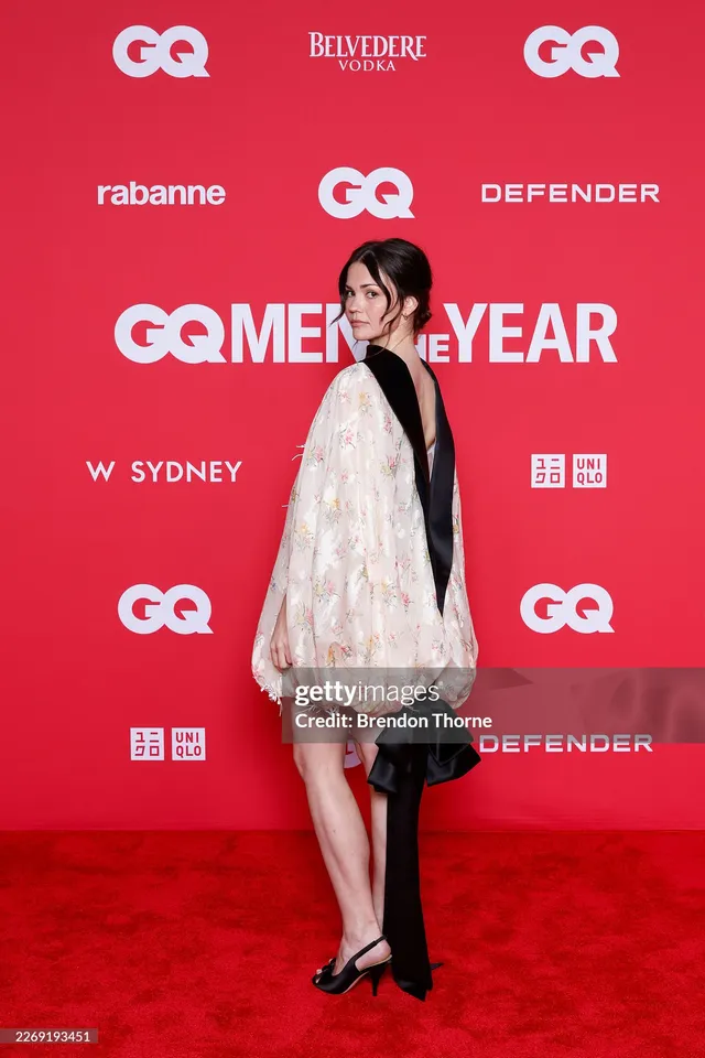 Maia Mitchell wearing a pale cape dress and heels on a red carpet.