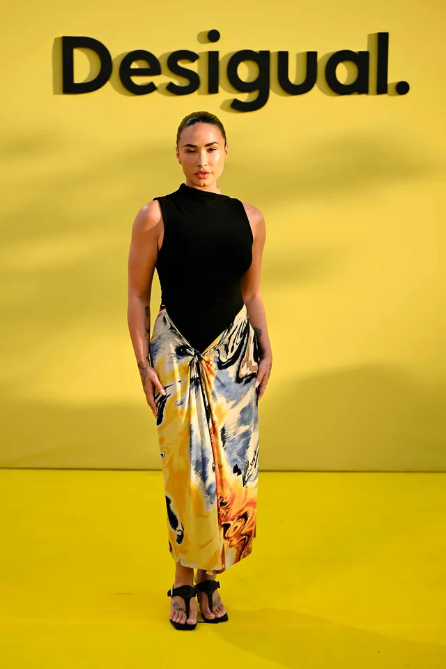 Demi Lovato posing against a yellow backdrop in patterned trousers and heels.