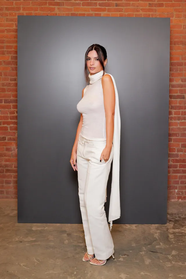 Emily Ratajkowski posing beside a brick wall in a pale outfit and heels.