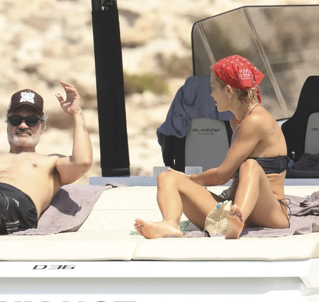 Rita Ora sitting barefoot on a beach lounger beside another adult.