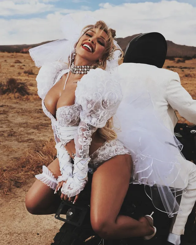 Doja Cat posing beside another person on a motorcycle in a white outfit and heels.