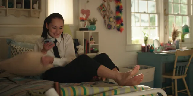 Lola Tung sitting on a bed with bare feet extended.