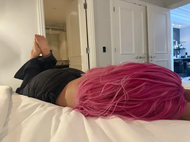 Bebe Rexha reclining on a bed with bare soles raised behind pink hair.