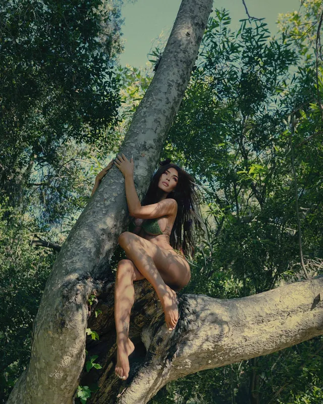 Megan Fox sitting on a tree branch outdoors.