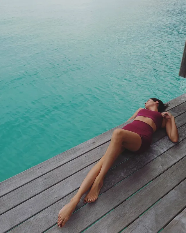 Sara Sampaio reclining barefoot on a wooden deck beside turquoise water.