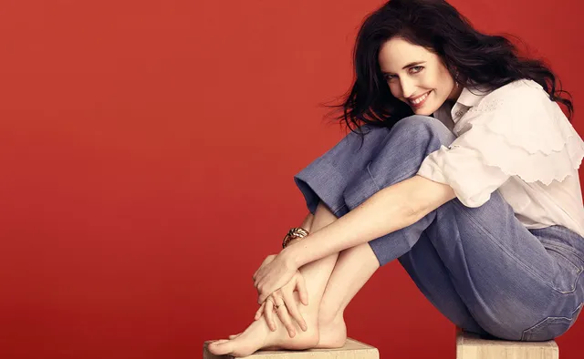 Eva Green seated against a red backdrop in a pale shirt and jeans.