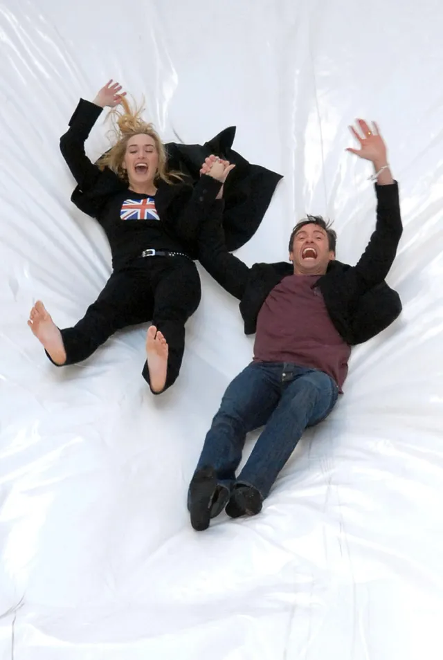 Kate Winslet reclining barefoot beside a companion on a white background.