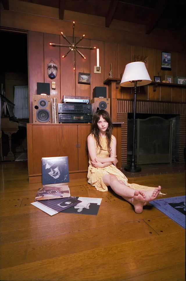 Sophie Thatcher sitting barefoot in front of a fireplace and stereo.