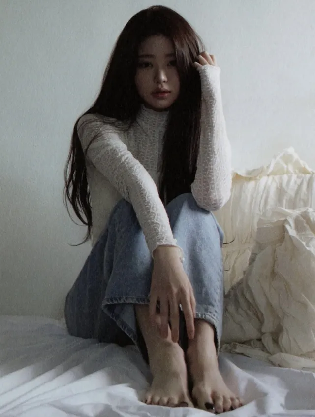 Jang Wonyoung sitting barefoot on a bed in jeans.