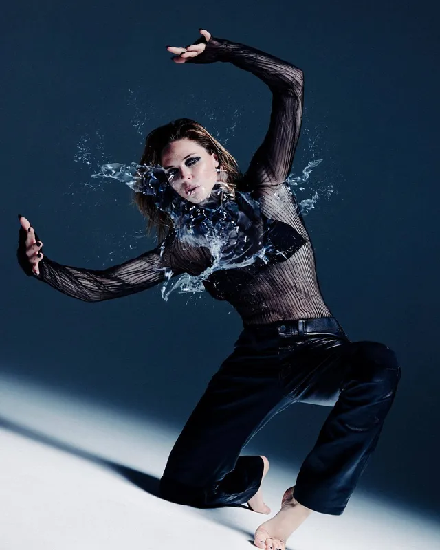 Rebecca Ferguson posing barefoot in dark trousers as water splashes across a studio set.