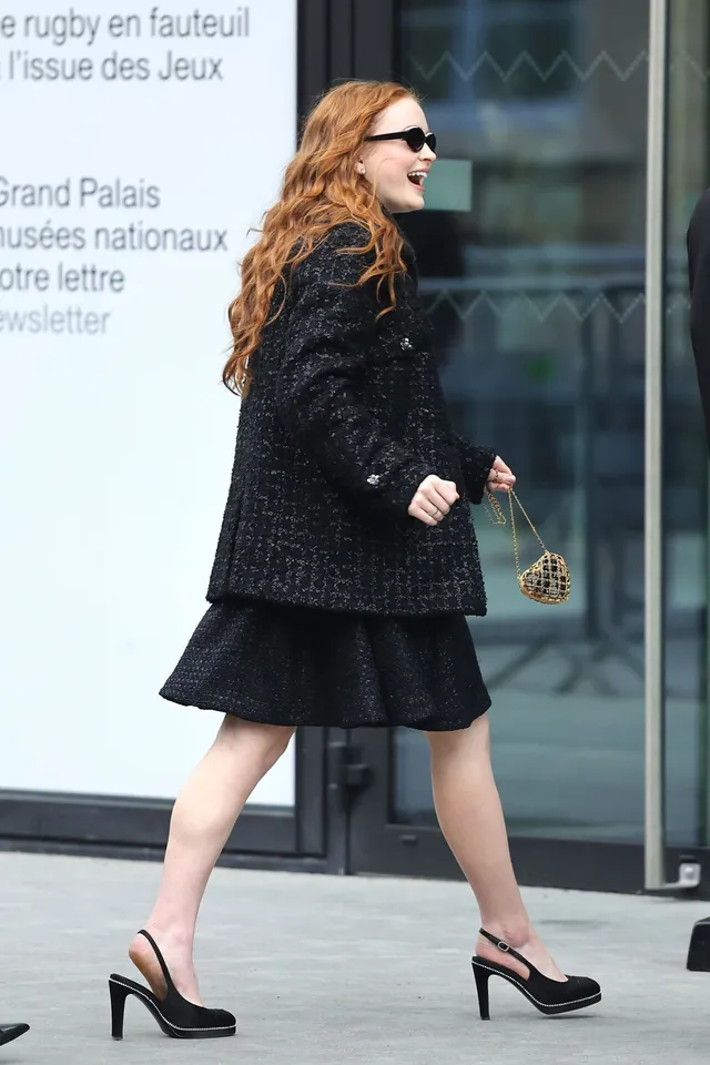Sadie Sink walking beside a building in a black dress and slingback heels.