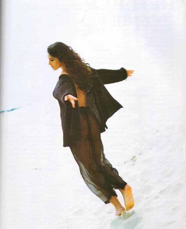 Monica Bellucci walking barefoot along a beach in a flowing dark outfit.