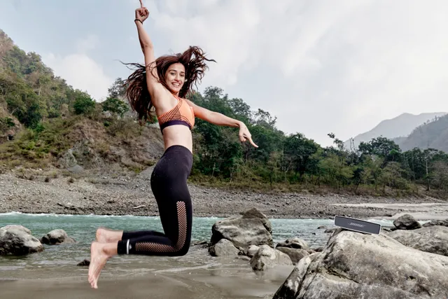 Disha Patani jumping barefoot on a rocky shore in a black exercise outfit.