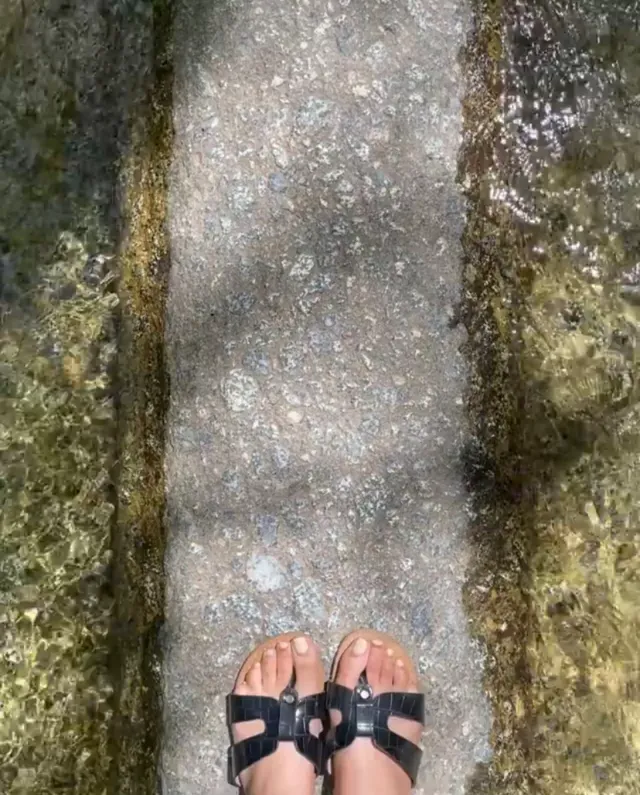 Emily Tosta wearing black sandals beside running water.