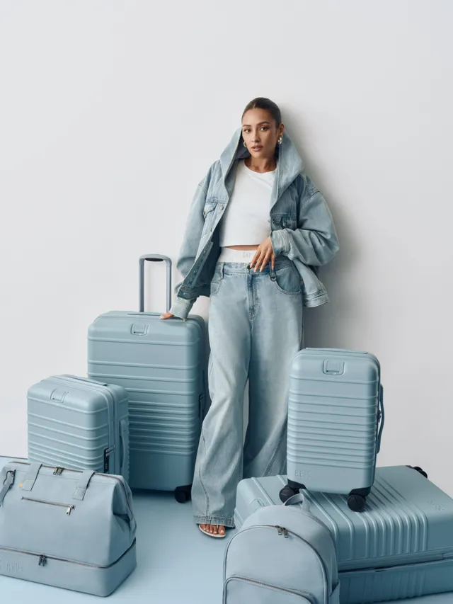 Shay Mitchell sitting beside matching luggage in a denim outfit.