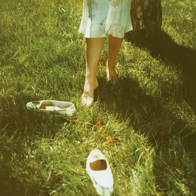 Lana Del Rey standing barefoot on grass beside a pair of pale shoes.