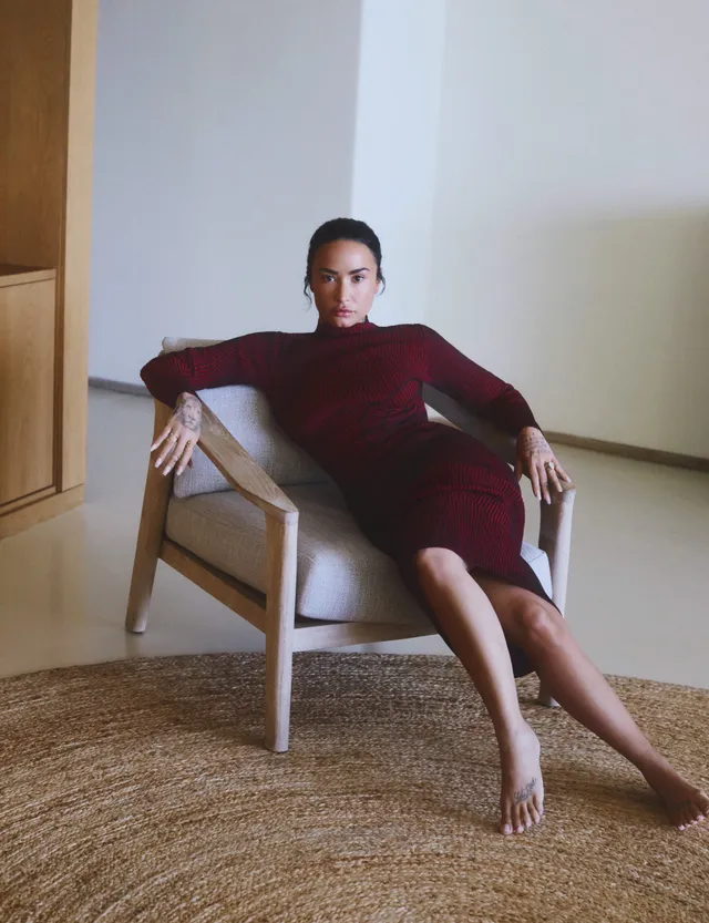 Demi Lovato sitting barefoot in an armchair in a burgundy dress.