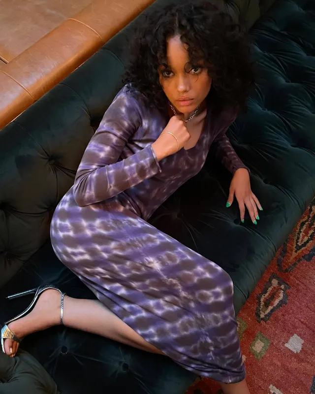 Whitney Peak reclining on a green sofa in a purple dress and heels.