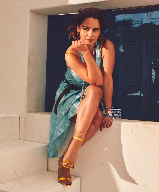 Emilia Clarke sitting on steps in a blue dress and gold heels.
