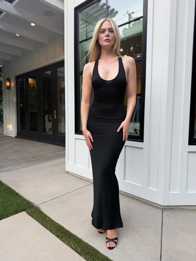 Elle Fanning posing outside in a black dress and heeled sandals.