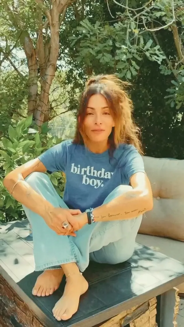 Sarah Shahi sitting on an outdoor bench in a blue T-shirt and jeans.