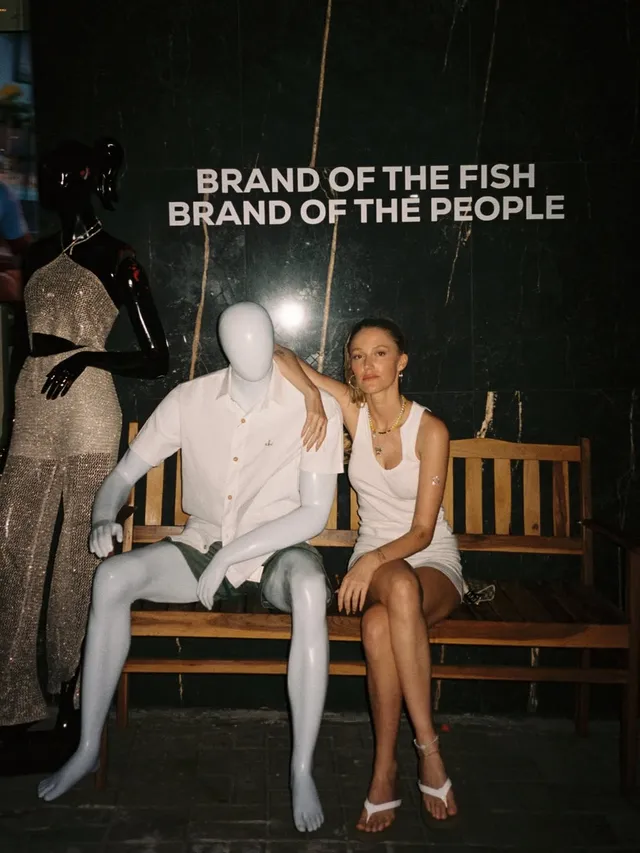 Maika Monroe sitting in white flip-flops beside a display.