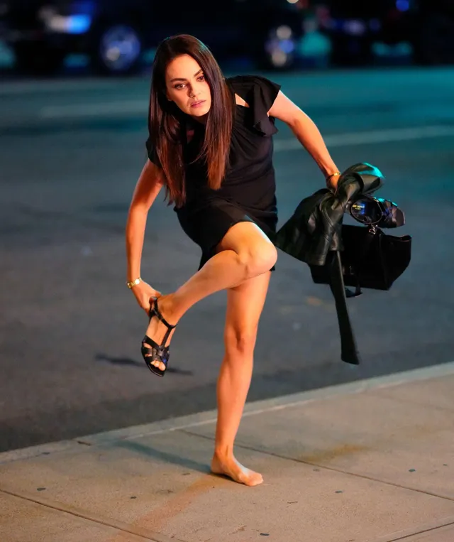 Mila Kunis standing in a black dress with one bare foot and one heeled sandal.