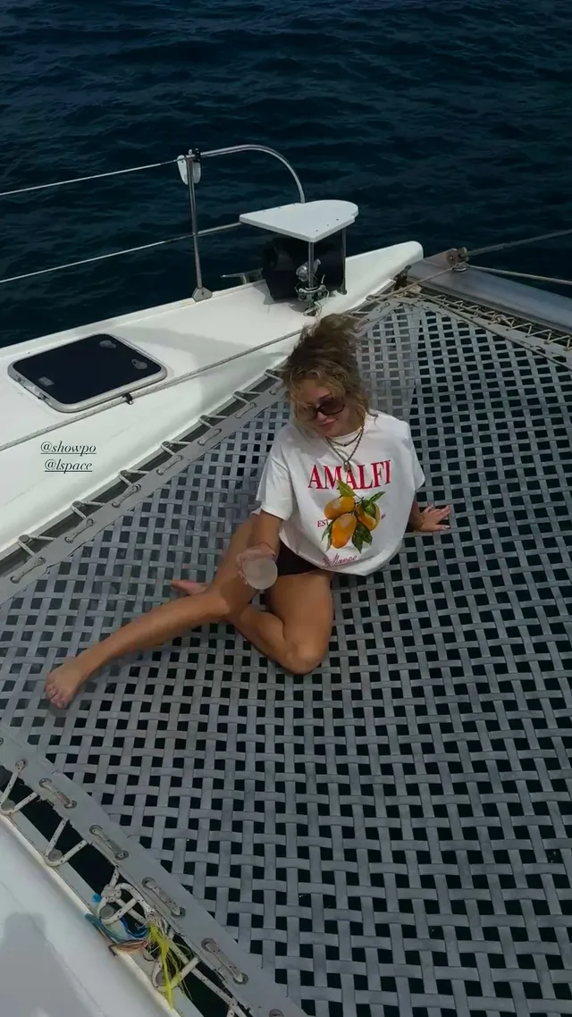 Brec Bassinger sitting barefoot on a catamaran net in a T-shirt and shorts.