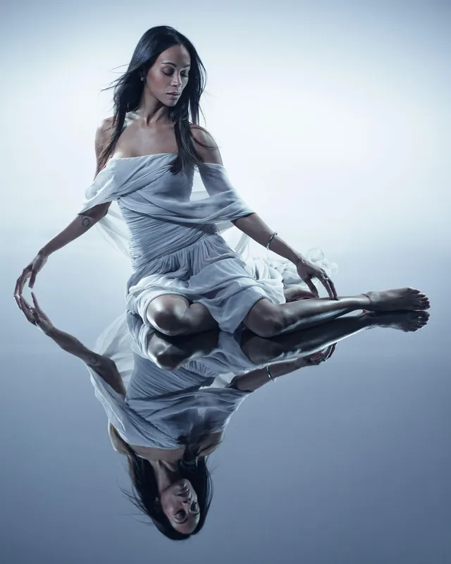 Zoe Saldaña sitting in a pale blue dress on a reflective surface.