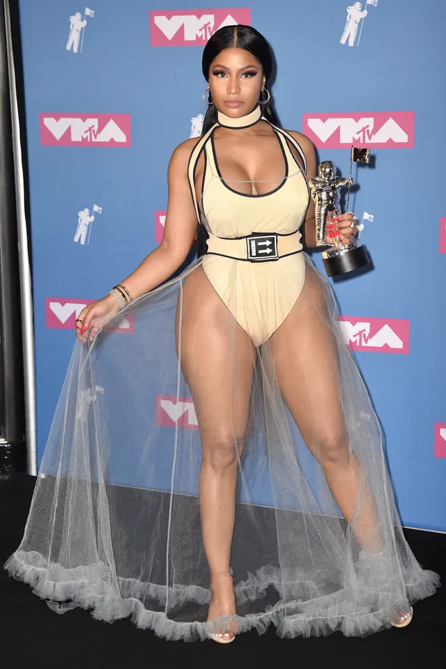 Nicki Minaj standing at an awards event in a pale outfit and sheer skirt.