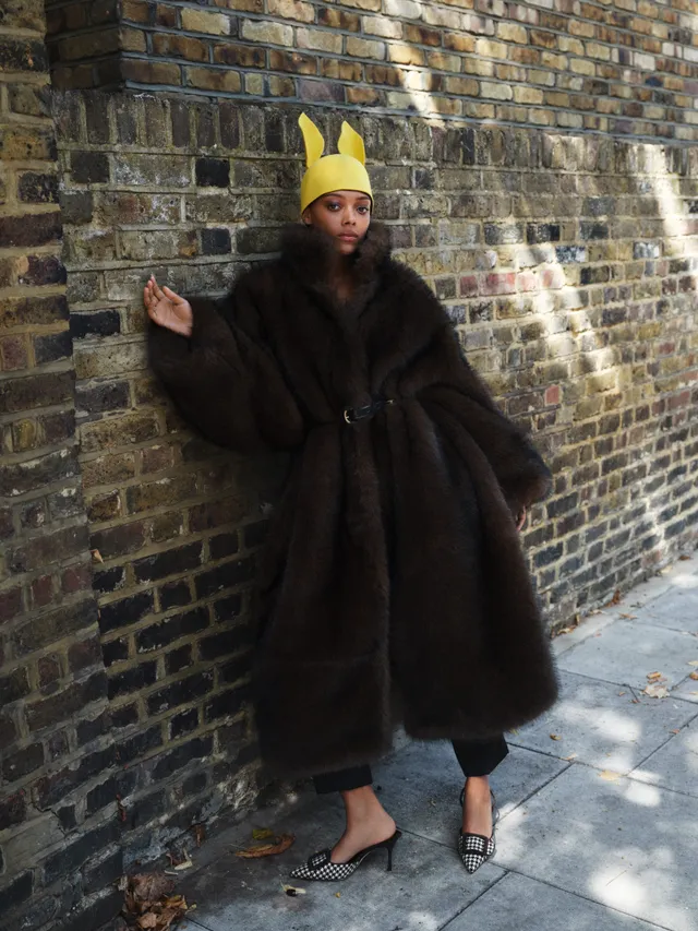 Sophie Wilde standing beside a brick wall in a dark coat and pointed heels.