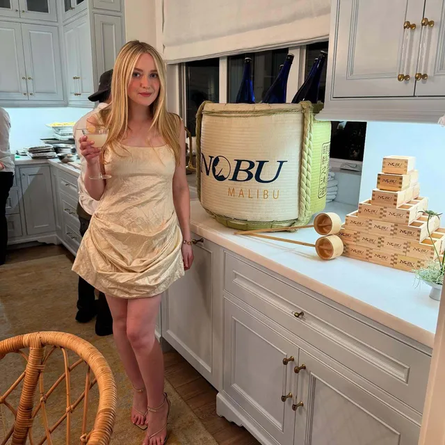 Dakota Fanning standing in a kitchen in a pale dress at a birthday celebration.