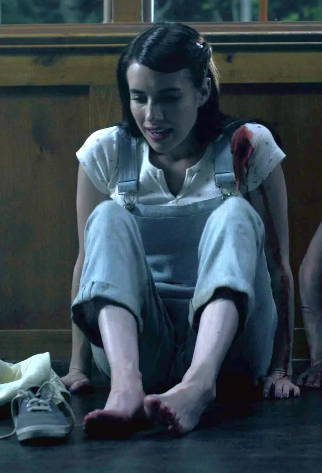 Emma Roberts sitting barefoot on the floor in gray overalls in a screen scene.