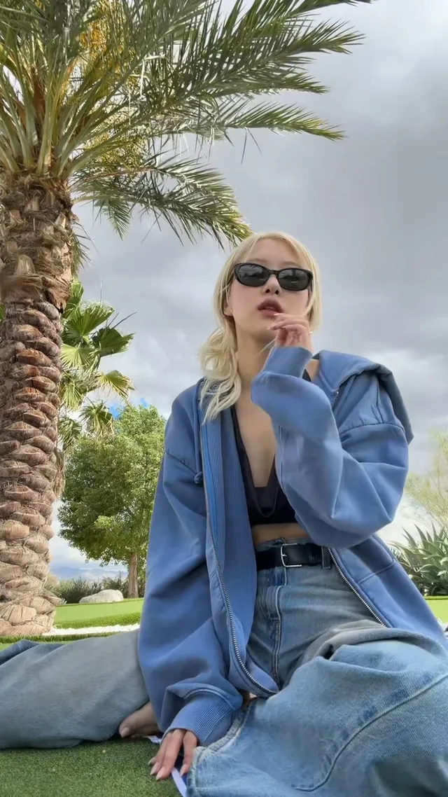 Rosé sitting on a lawn in a blue jacket, jeans and sunglasses.