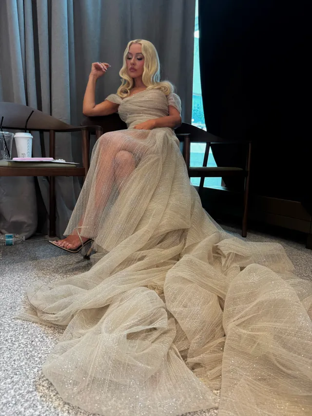 Christina Aguilera sitting in a long pale gown and metallic heels.