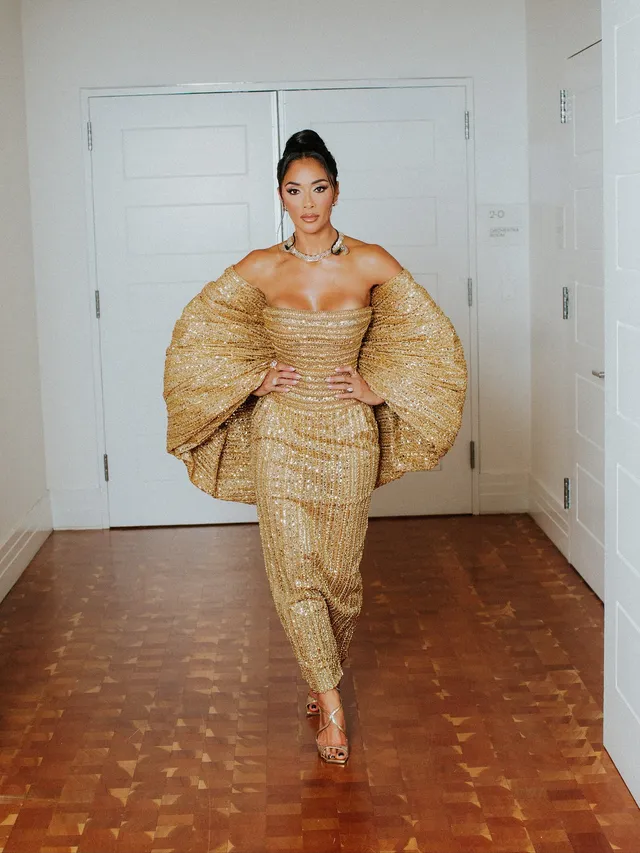 Nicole Scherzinger standing indoors in a gold gown and metallic heels.