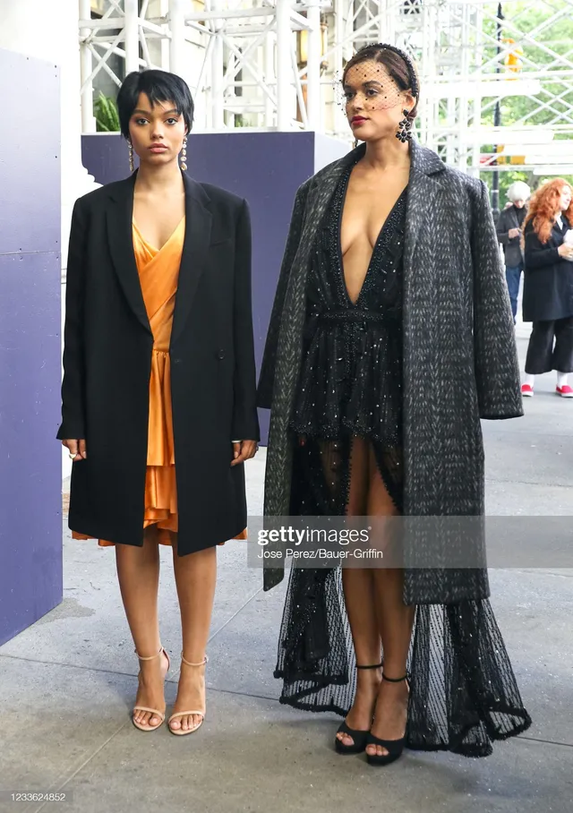 Whitney Peak posing beside a guest in an orange dress, black coat and heels.