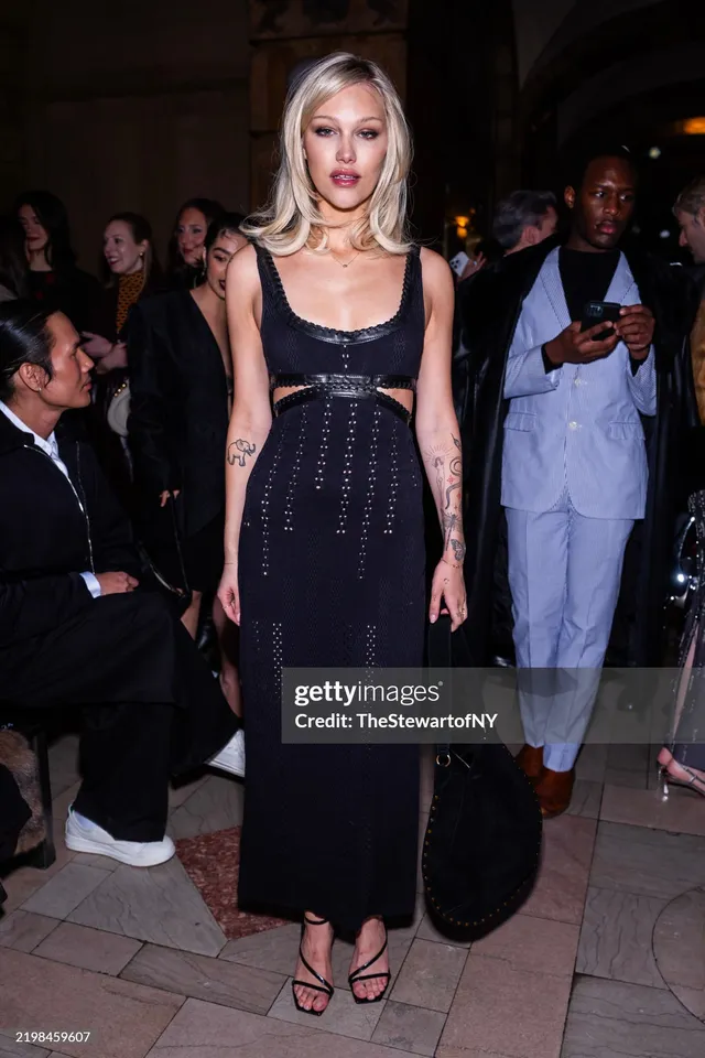Grace VanderWaal standing at a fashion event in a black dress and heels.