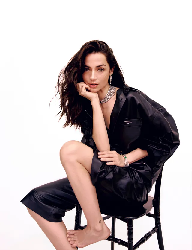 Ana de Armas seated barefoot on a stool in a black outfit.