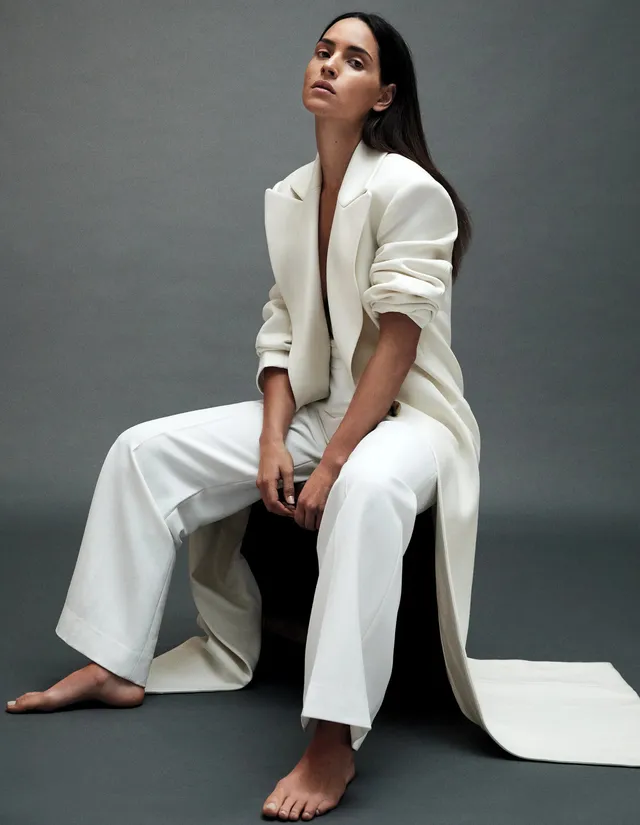 Adria Arjona posing barefoot in a white suit on a studio stool.