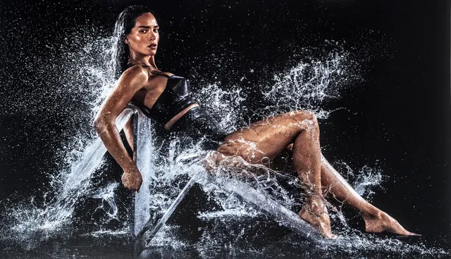 Adria Arjona posing barefoot in a black dress surrounded by splashing water.