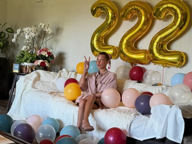 Bella Hadid sitting barefoot on a bed beside birthday balloons.
