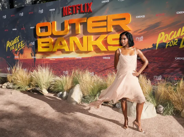 Carlacia Grant posing in a pale dress and heels before a premiere backdrop.