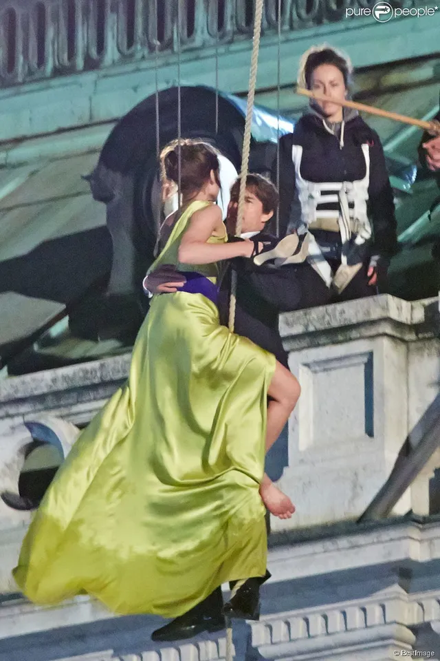 Rebecca Ferguson sitting barefoot on a rooftop railing in a yellow dress beside a companion.