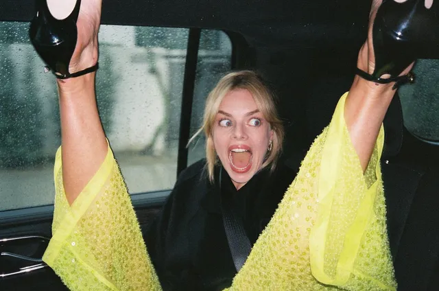 Samara Weaving raising black heels while sitting inside a vehicle.