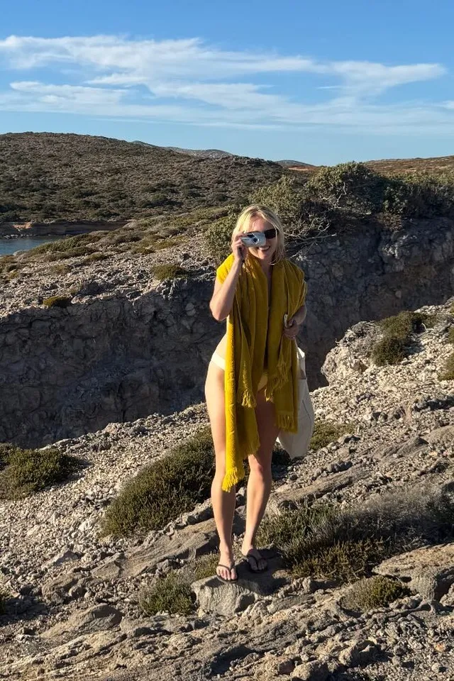 AnnaSophia Robb standing on rocks beside the coast in a yellow dress.