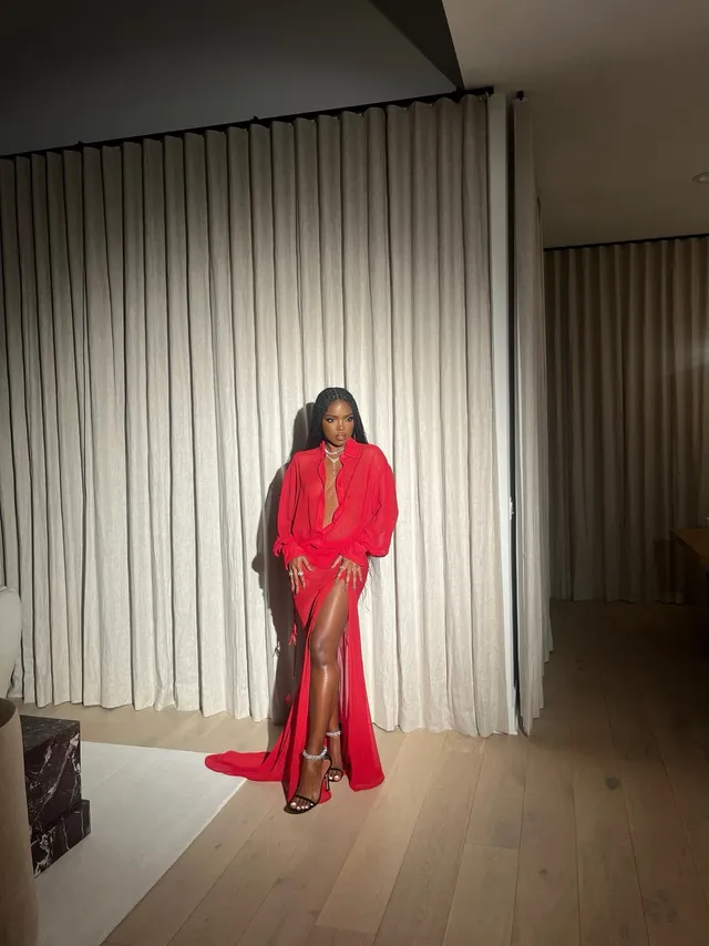 Ryan Destiny standing indoors in a long red gown and metallic heels.