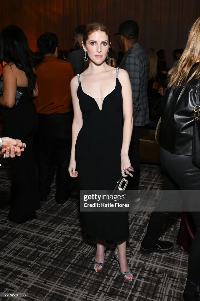 Anna Kendrick posing at an event in a black dress and heels.