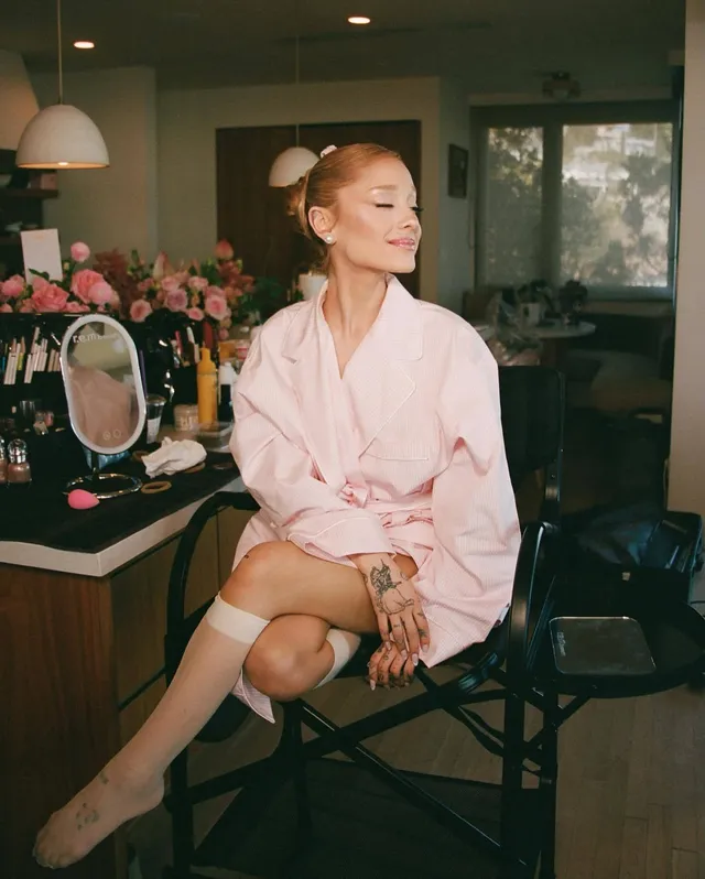Ariana Grande: seated barefoot in a dressing-room chair wearing a pink outfit.