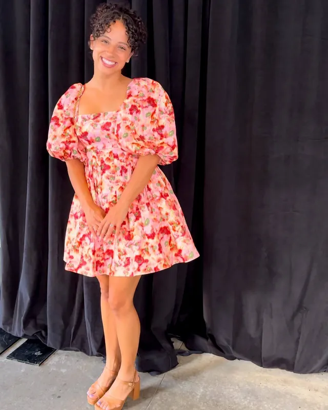 Kiana Madeira standing in a floral dress and pale heels before a curtain.